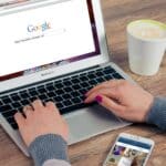 Hands typing on MacBook Air with Google search open, coffee nearby.
